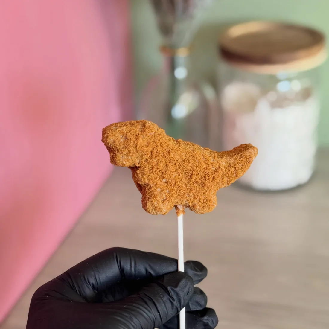 Gloved hand holding a dinosaur nugget shaped cake pop against a pink wall