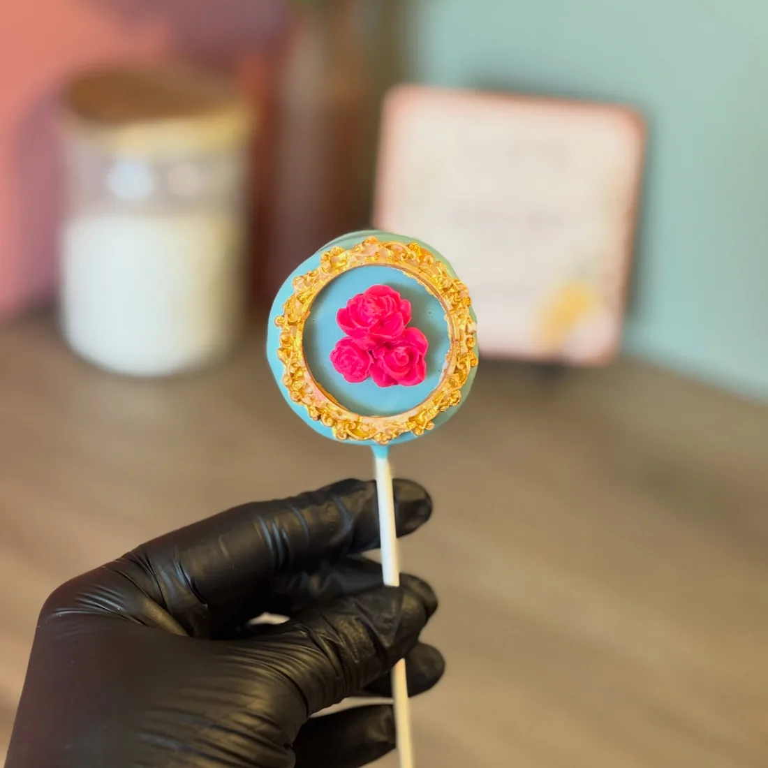 Round cake pop with a teal centre, pink piped roses and a gold crumb rim, held in a gloved hand