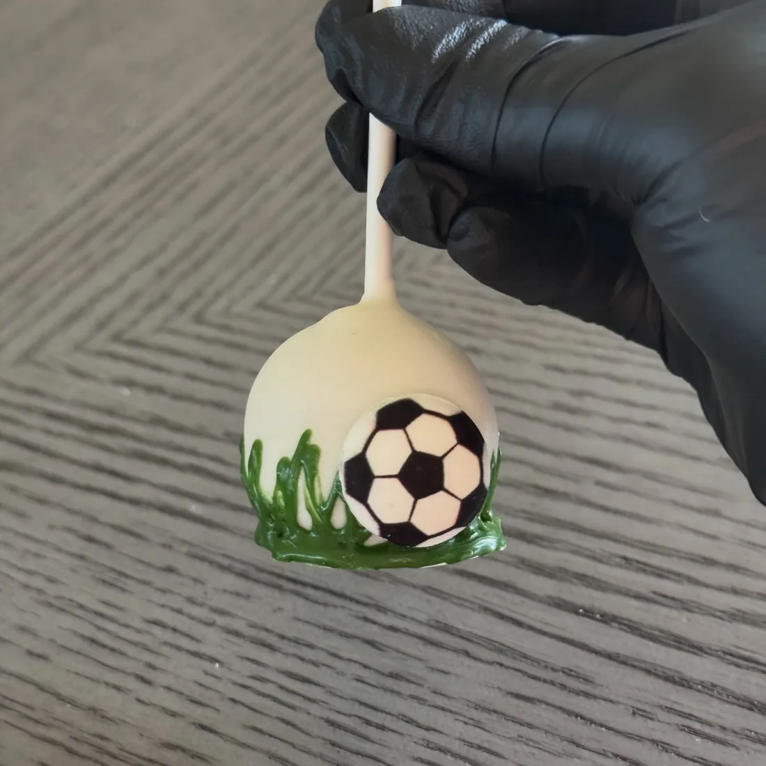 Gloved hand holding a white cake pop with a soccer ball and piped green grass