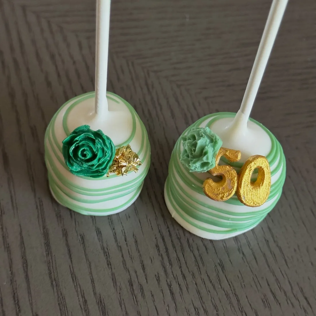 Two green marbled cake pops, one with an emerald piped rose and one with a gold 50