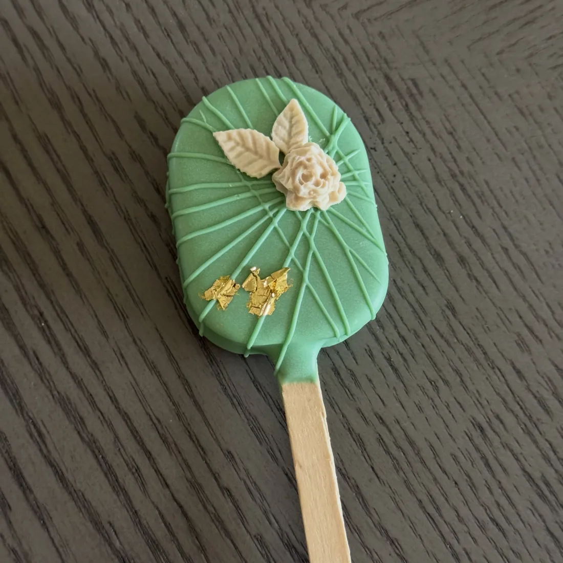 Emerald green cakesicle on a stick with a white piped rose, leaves and gold leaf flakes