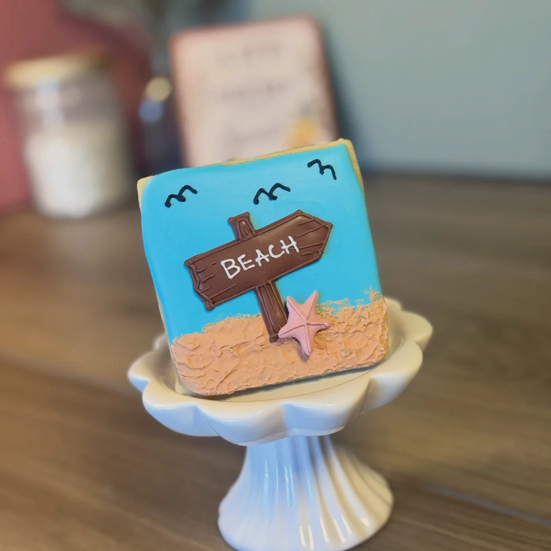 Square cookie decorated with a wooden beach signpost, sand, a pink starfish and seagulls in a blue sky