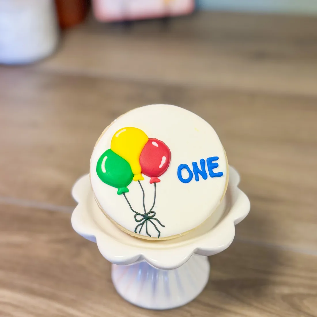 Round iced sugar cookie on a white pedestal stand, decorated with green, yellow and red balloons and the word ONE in blue