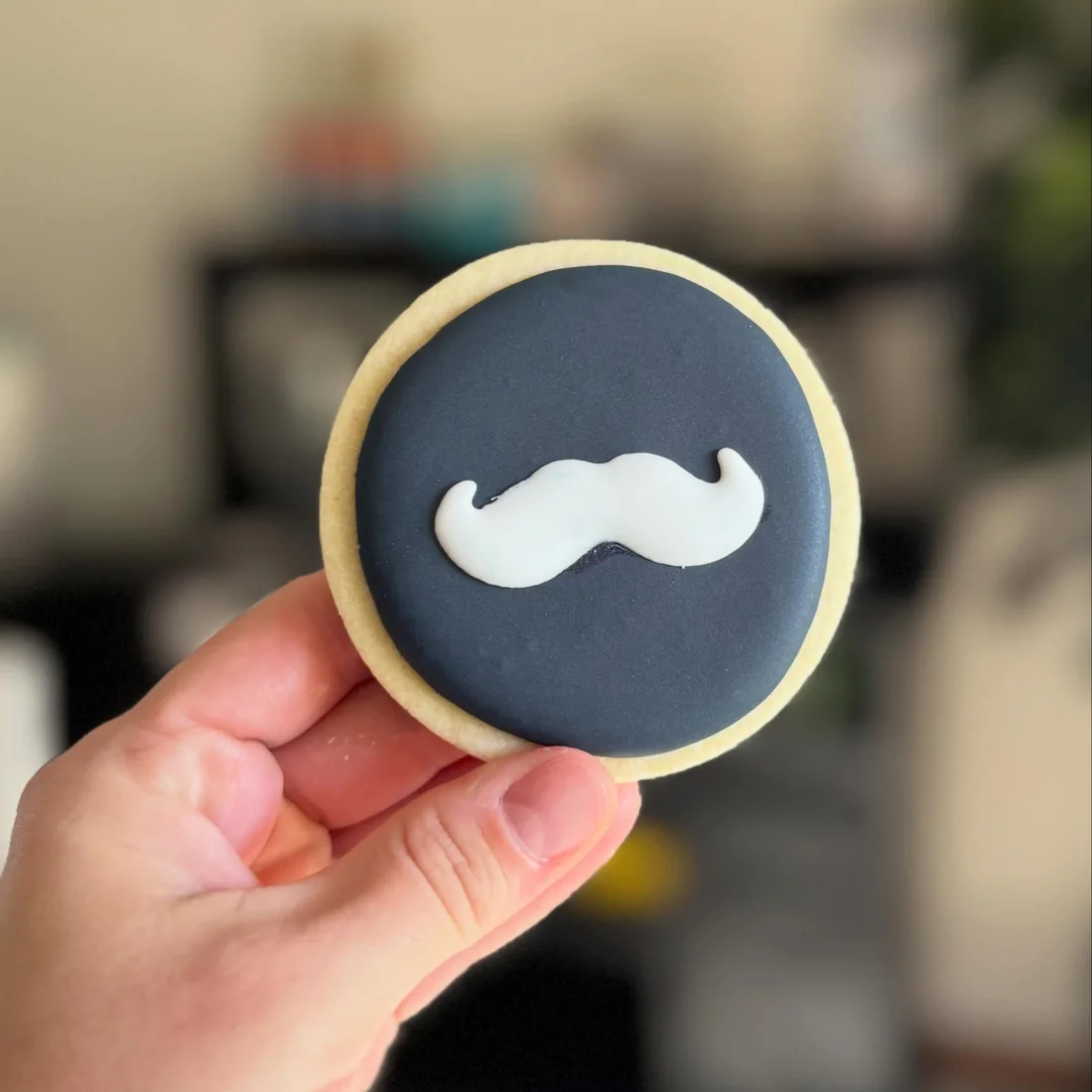 Hand holding a navy round cookie with a white royal icing mustache
