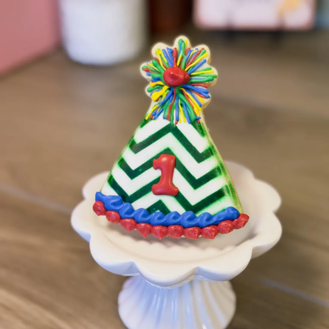 Party hat shaped sugar cookie on a white pedestal stand, iced with a green chevron pattern, a red number one and a multicoloured pom-pom