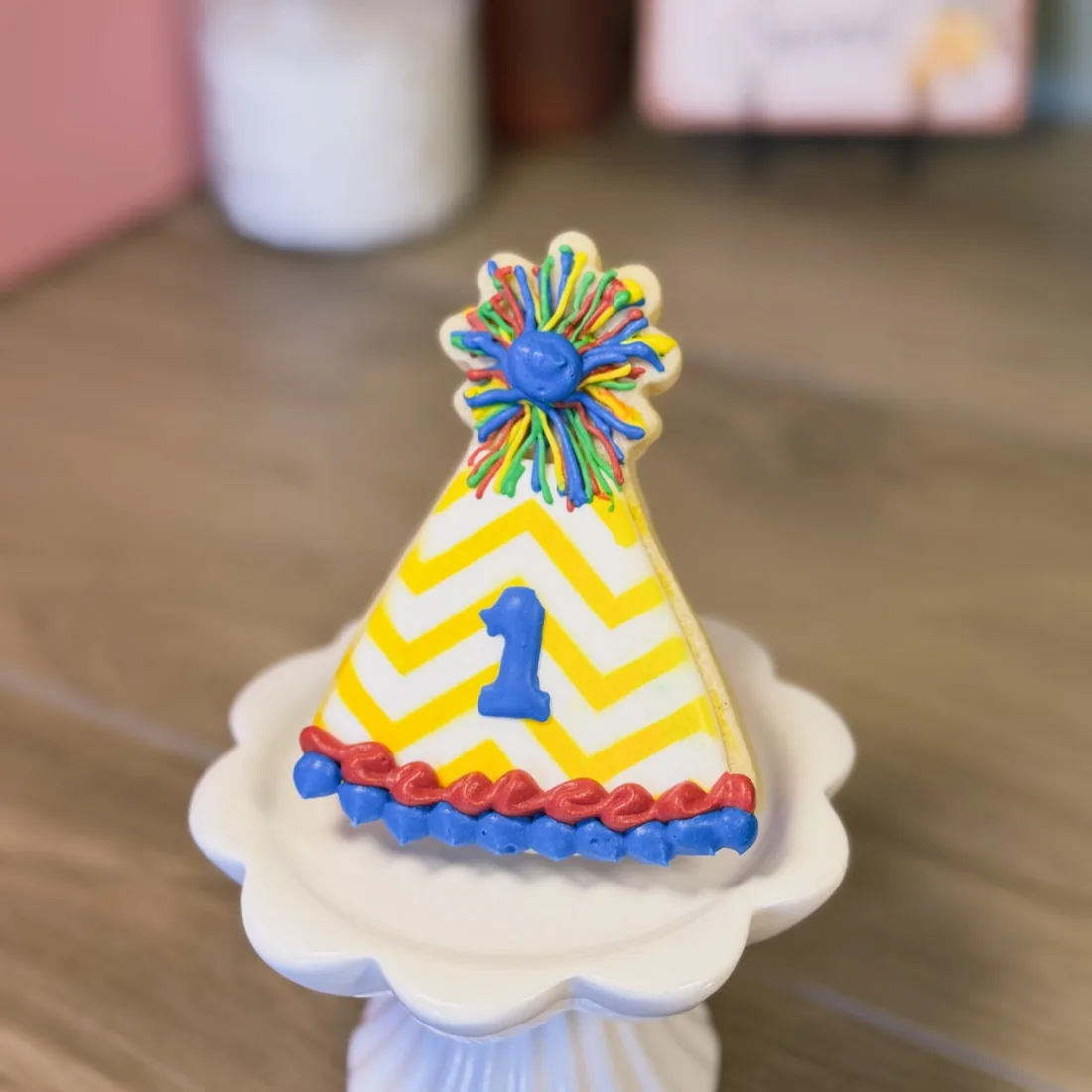 Party hat shaped sugar cookie on a white pedestal stand, iced with a yellow chevron pattern, a blue number one and a multicoloured pom-pom