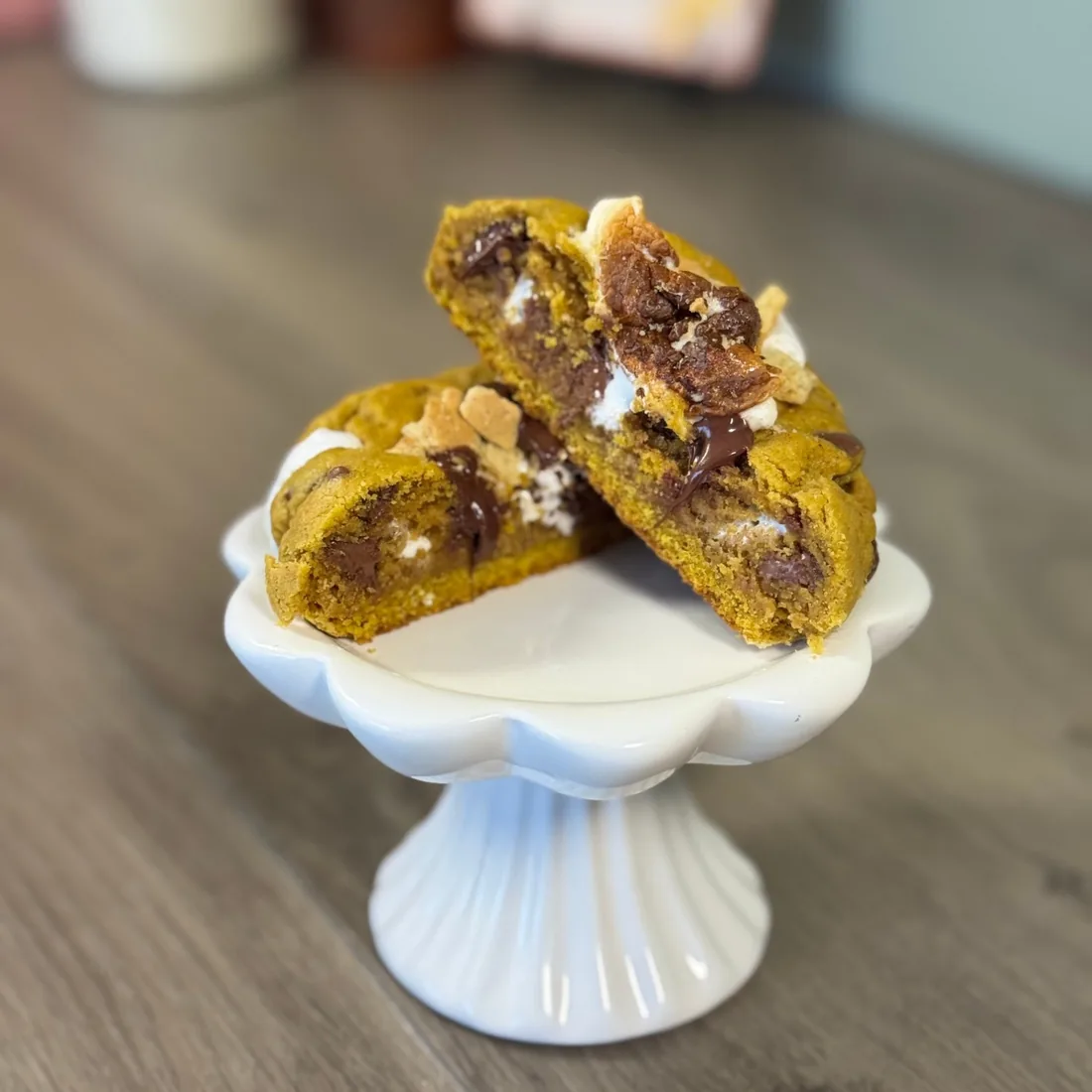 Two halves of a pumpkin s'more cookie stacked on a white pedestal stand, showing melted chocolate, marshmallow and graham cracker pieces