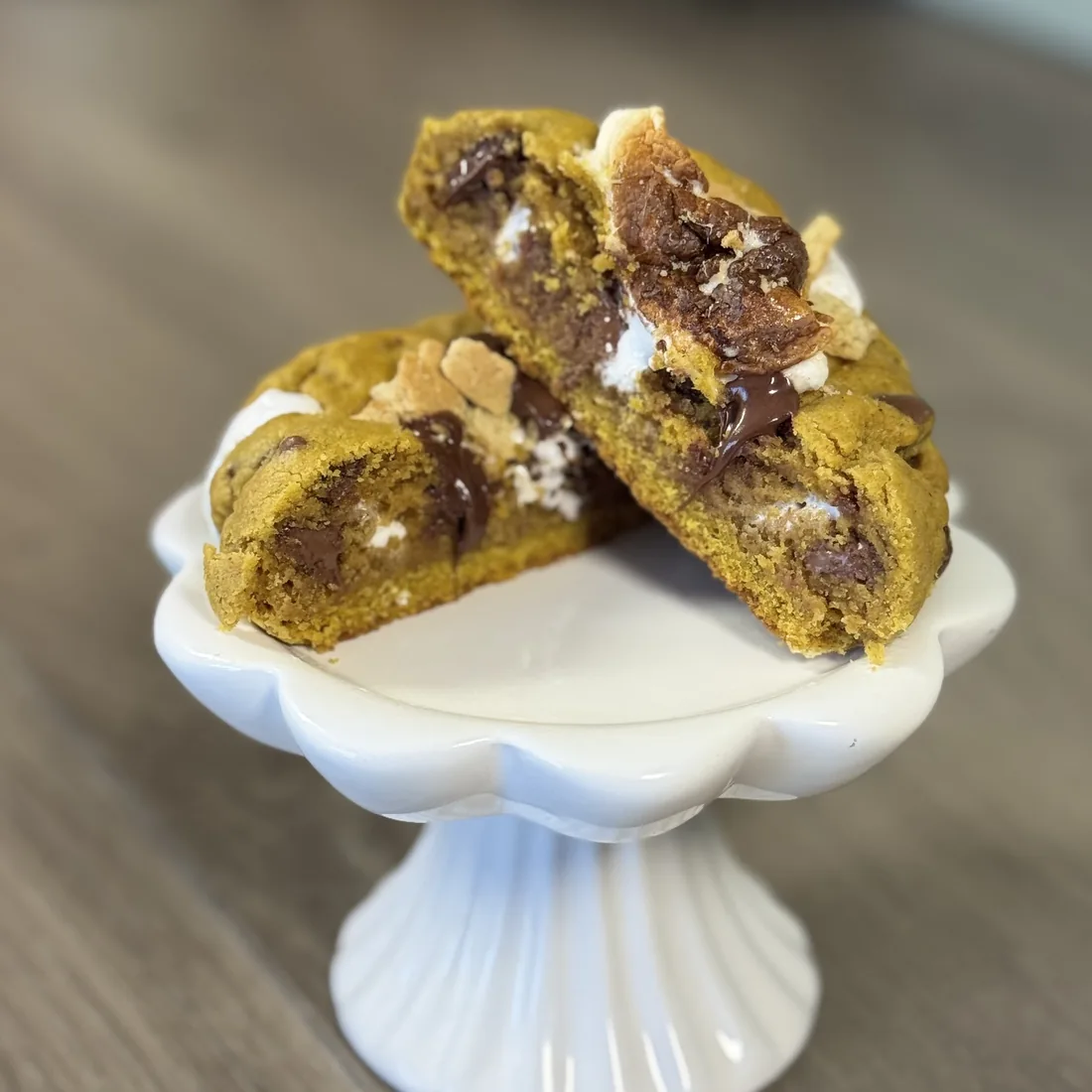 Pumpkin s'more cookie split in half on a white pedestal stand, showing a gooey marshmallow center with melted chocolate and graham cracker pieces on top