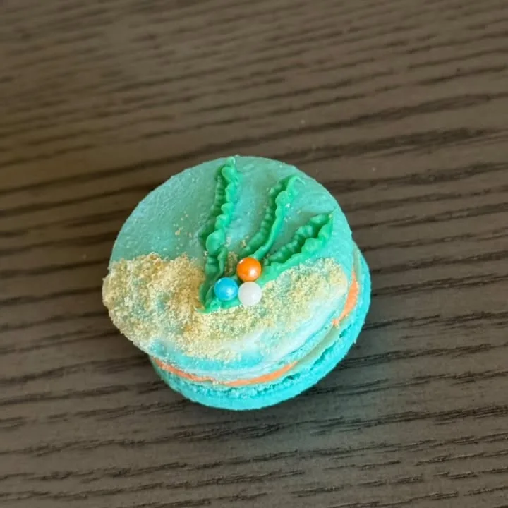 Teal macaron piped with green coral over a sandy crumble edge with sugar pearls
