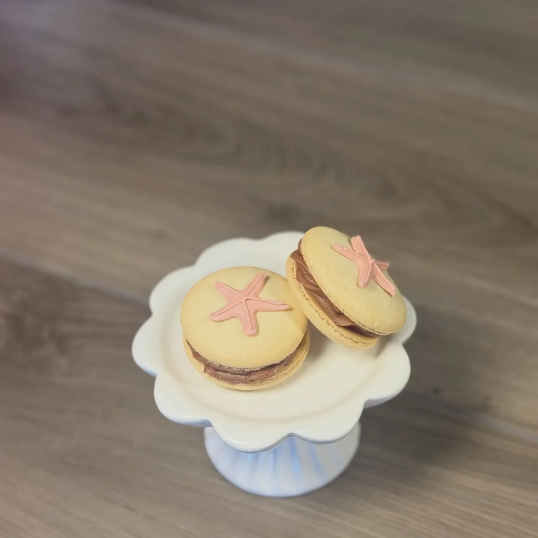 Two pale vanilla macarons topped with pink fondant starfish on a scalloped cake stand