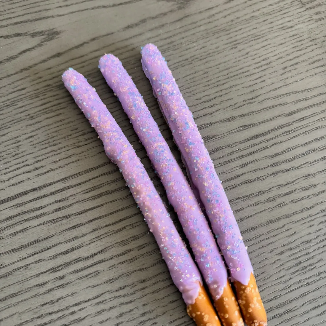 Three pretzel rods dipped in lavender white chocolate with iridescent sugar crystals
