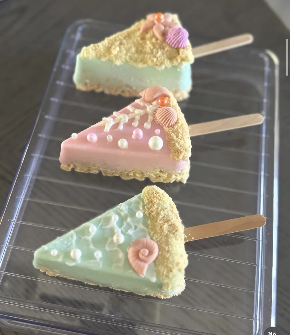 Three beach Rice Krispie treats in mint and pink with shells and sandy crumble on a tray