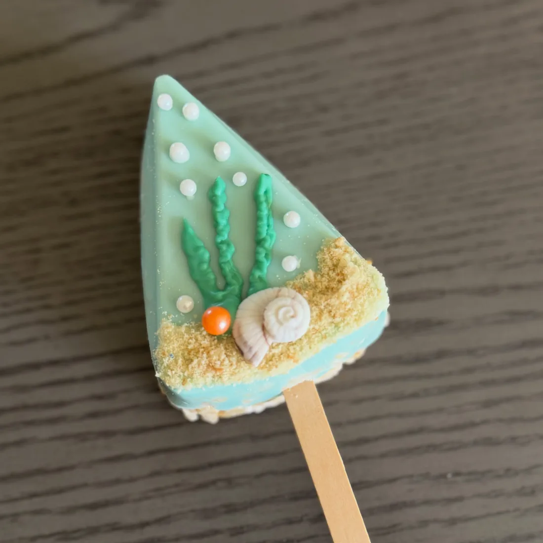 Mint green triangle Rice Krispie treat with piped seaweed, sugar pearls, a snail shell and sandy crumble